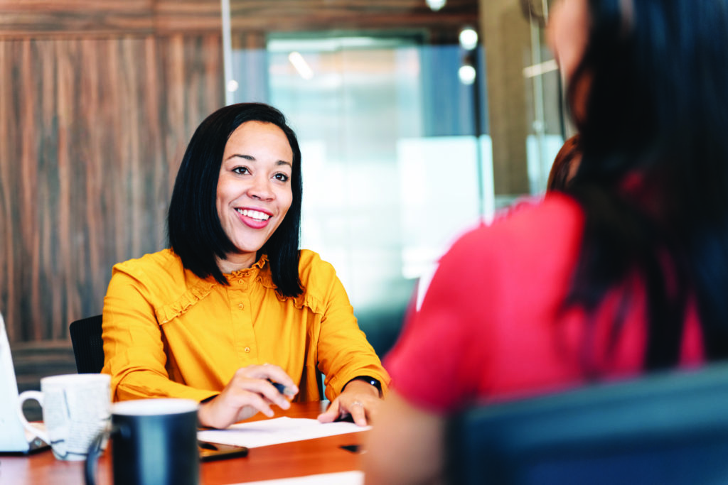 A woman sits across a desk from another woman. She is smiling and wearing an orange shirt.