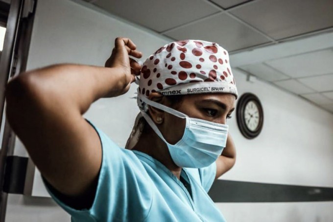 Female nurse wearing green scrubs is tying a mask around her hair cover