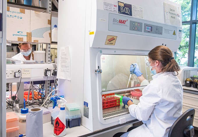 Researcher in a lab is using a syringe to fill a beaker on a counter. She is wearing a white lab coat and gloves