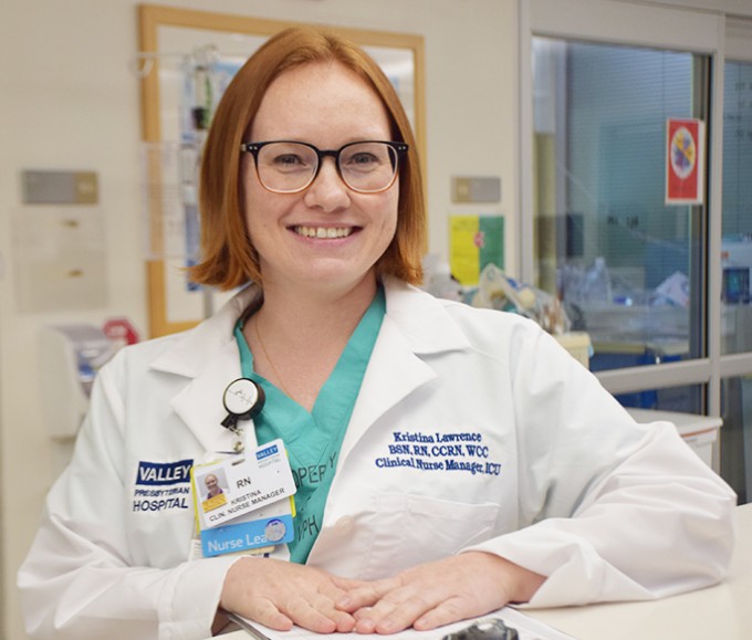 Registered Nurse Kristina Lawrence wearing scrubs and a white coat stands smiling with her unit in the background