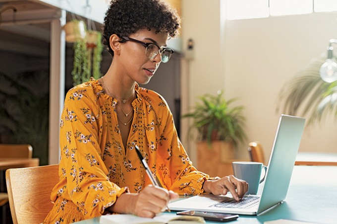 A nurse in casual clothing is sitting at a desk typing on her computer and writing things down