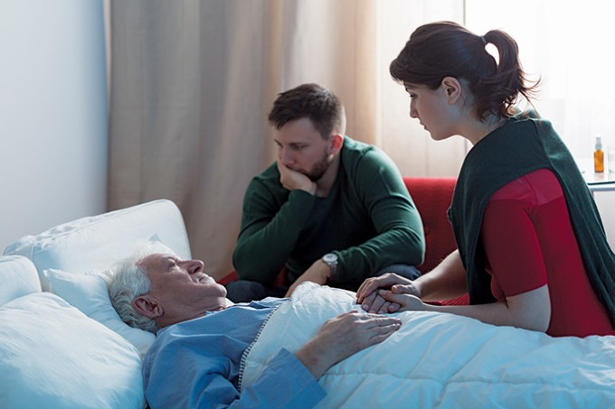 A woman and man sit at the bedside of an older man. They are holding his hands and look sad
