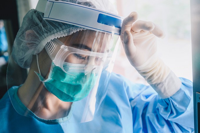 A nurse in scrubs, mask, goggles, and gloves is leaning against her hand looking tired