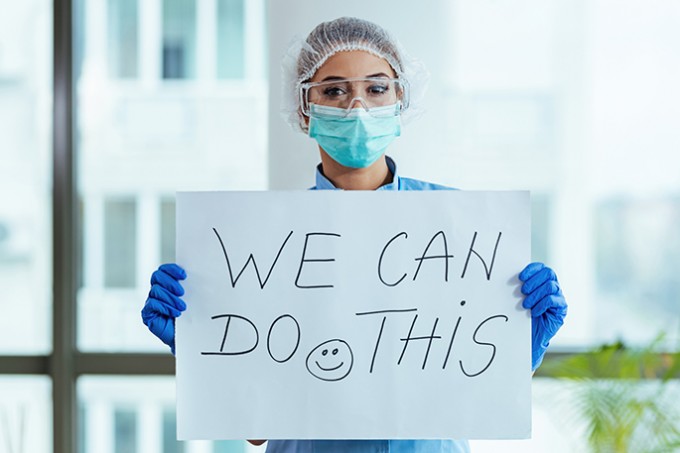 A nurse in PPE is holding a sign that says "We Can Do This" with a smile on the sign
