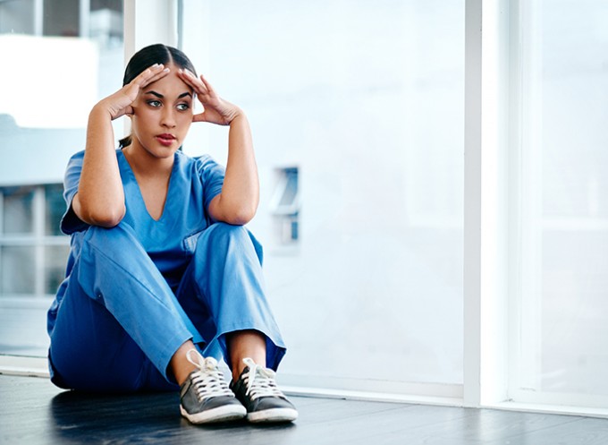 A nurse in scrubs is sitting on the hospital floor with her arms on her knees and her head in her hands.