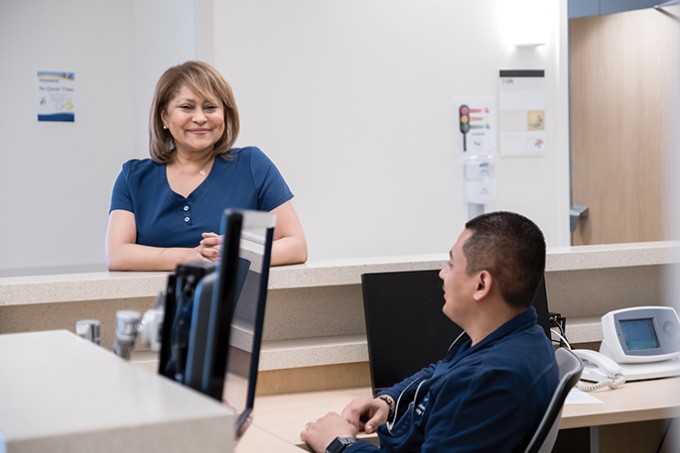 Registered Nurse Rita Calderon stands smiling to her college that is sitting in the foreground on a chair by a computer.