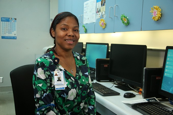 Registered Nurse Michelle McKoy is wearing a green blouse and her badge. She smiles as she sits by a computer.