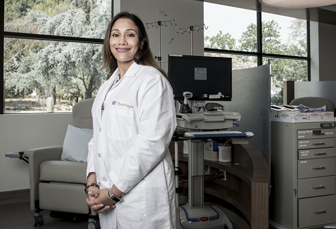 Registered Nurse Anita Karra is wearing a white coat and smiling towards the camera with a patient room behind her.