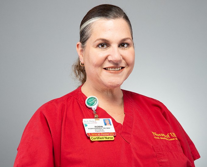 Registered Nurse Robin Krasner wears red scrubs and is standing and smiling in front of a gray background
