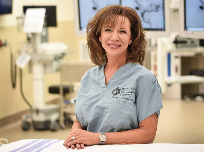 Registered Nurse Bonita Aceto stands smiling in blue scrubs with the operating room in the background.