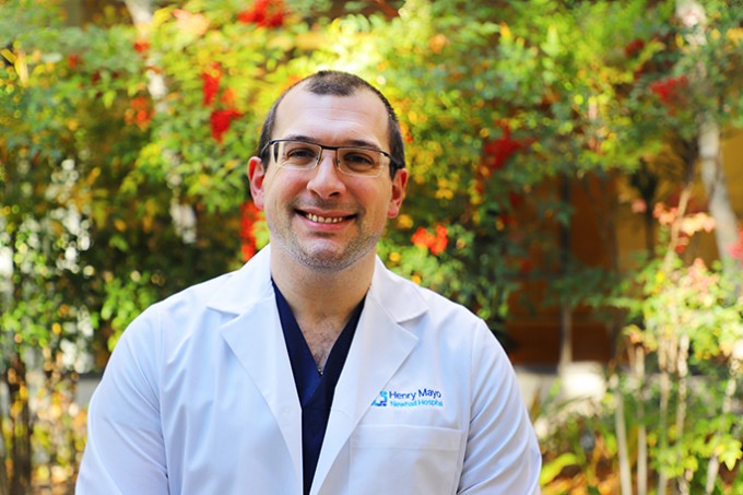 Registered Nurse Michael Russo stands smiling in a white coat and scrubs, with green bushes with red flowers in the background.