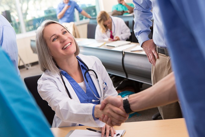 A nurse is smiling seated at a desk and shaking the hand of another nurse who is standing in front of her