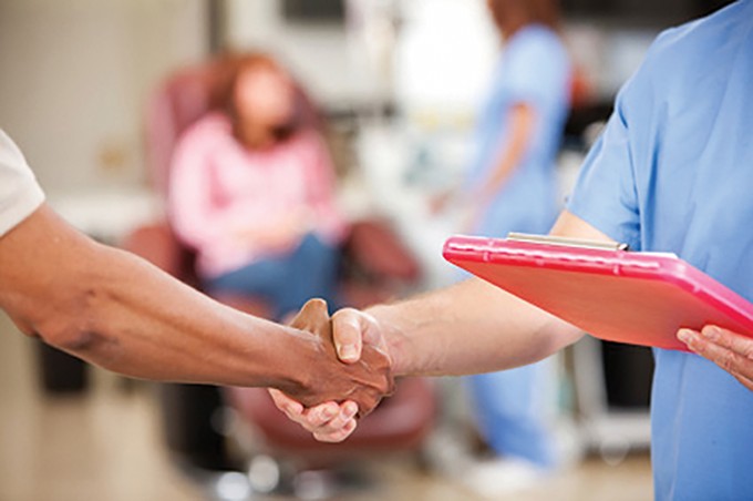 A nurse in scrubs is shaking hands with someone else. The nurse is holding a pink clipboard in their other hand