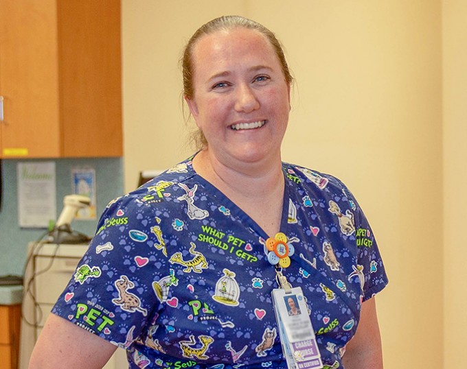 Registered Nurse Tami Hitchcock stands smiling in colorful scrubs. She is standing in front of a yellow wall.