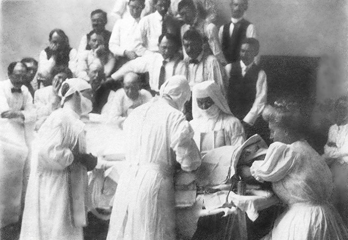 Alice Magaw working with doctors to take care of a patient. They are all wearing white gowns and head covers with masks on
