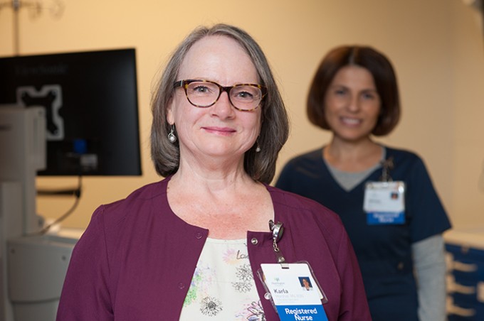 Registered Nurse Karla Marshall stands smiling in a blouse and sweater with her badge on, and a coworker on the computer behind her.