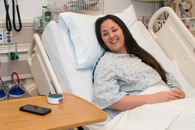 Female patient lying in a hospital bed and wearing a hospital gown, is talking to a voice assistant on the table next to her