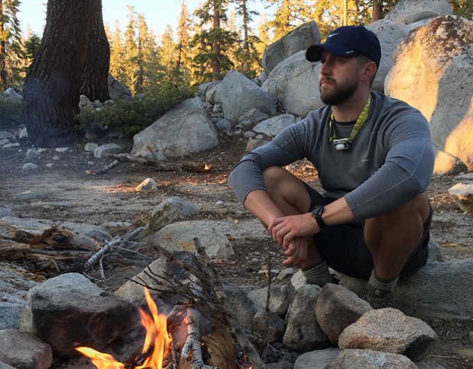 Jon Garrison sits aside a fire in the wilderness. Rocks and trees surround him.