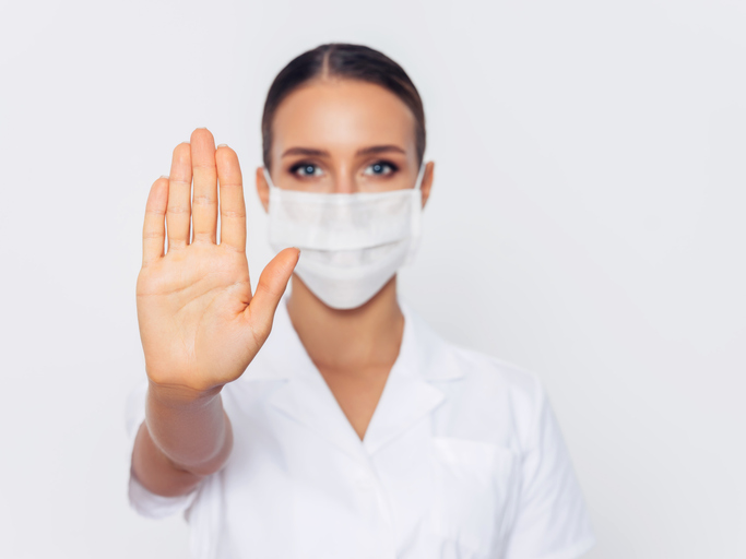 A nurse in white scrubs and wearing a white mask is holding up her hand in a stop position