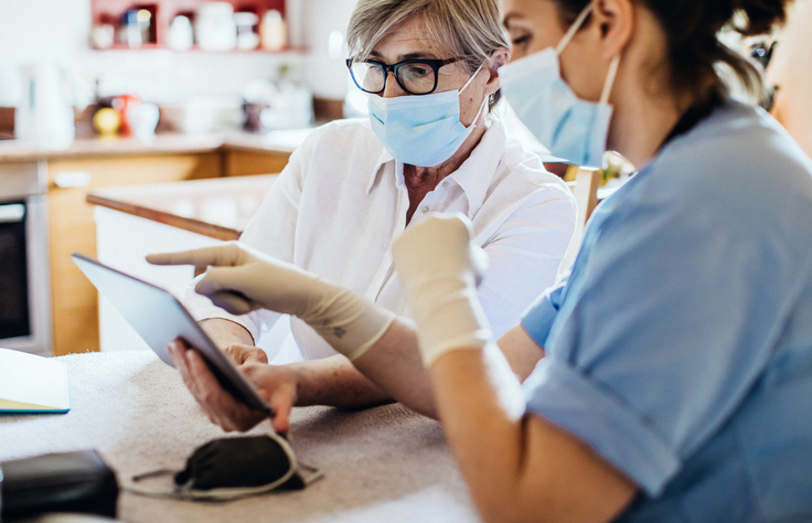 Two nurses, one with scrubs on and the other wearing a white coat, are discussing how to use a tablet that's in front of them