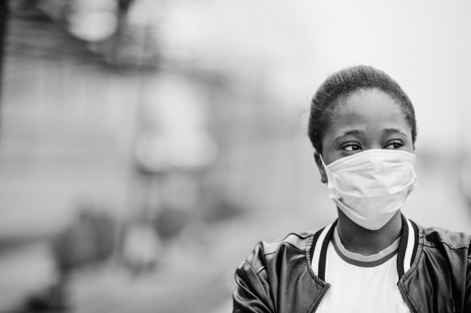 Black woman wearing a mask and looking to the right of the camera. The background is blurry and the whole photo is in black and white