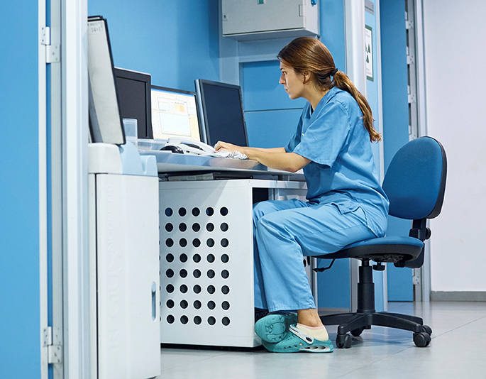 A nurse in scrubs is sitting on a chair in front of the computer that she is typing on