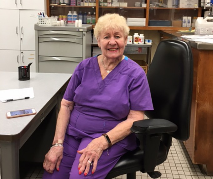 Registered Nurse Marti Sillman sits on an office chair in purple scrubs smiling at the camera