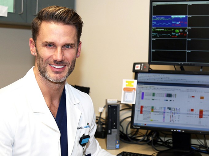 Registered Nurse Donovan Stewart is smiling in front of a computer while wearing a white coat and scrubs.