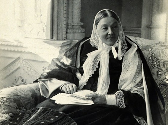 Florence Nightingale sits on a couch smiling in multiple layers of clothing