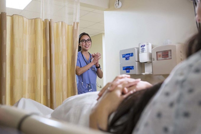 A patient sits on a bed while a nurse in scrubs is rubbing hand sanitizer on her hands by a curtain