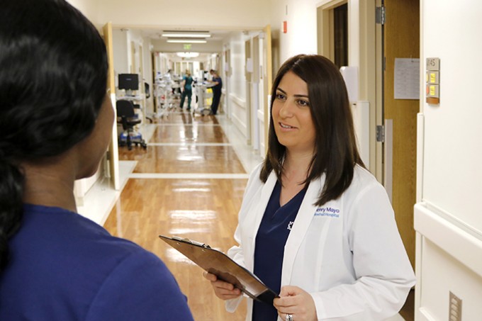 Registered Nurse Babasi Maradkel stands in a hallway with a white coat on. She is holding a clipboard and smiling, while talking to other staff members.