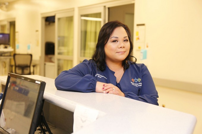 Registered Nurse Lyrose Ortiz stands smiling in blue scrubs, arms resting on a counter.