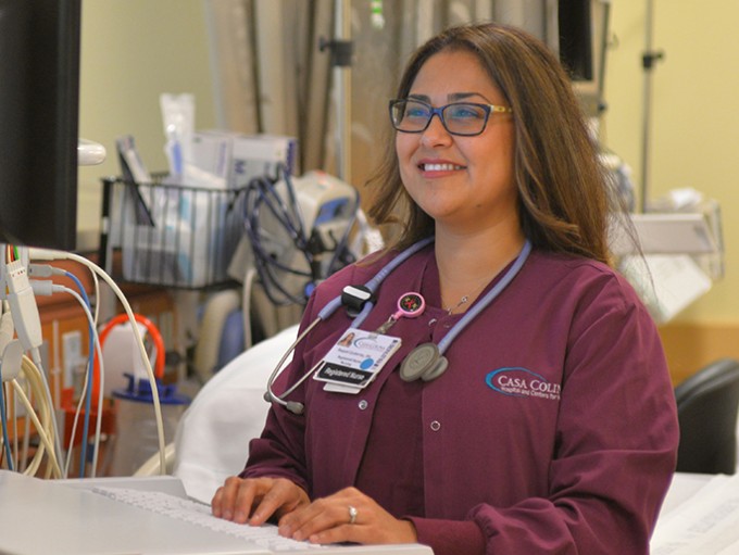 Registered Nurse Raquel Gutierrez is sitting in maroon scrubs, and is on the computer.