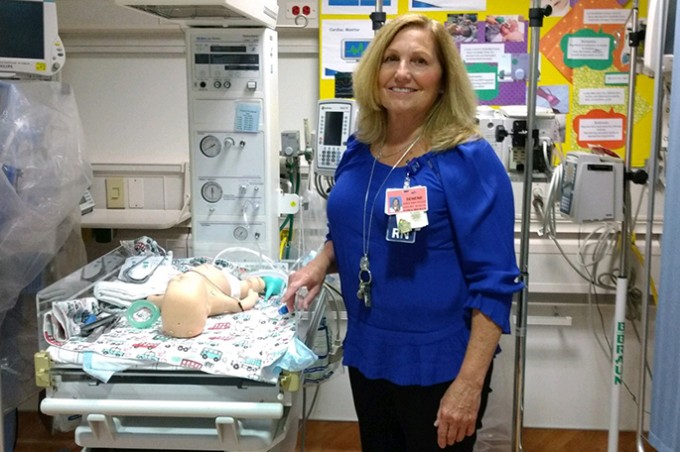 Registered Nurse Serene Owen stands smiling while holding a baby simulator that is resting on a table next to her.