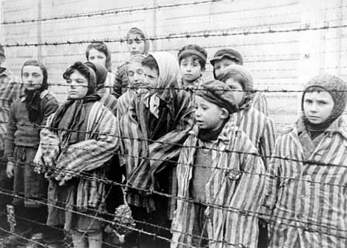 Groups of people in a concentration camp are behind barbed wire fencing and wearing striped uniforms