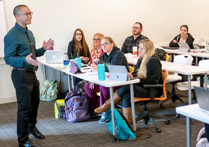 Edmund Alfonso stands in front of a group of students. He is wearing a button down, tie, and black pants, and is teaching.