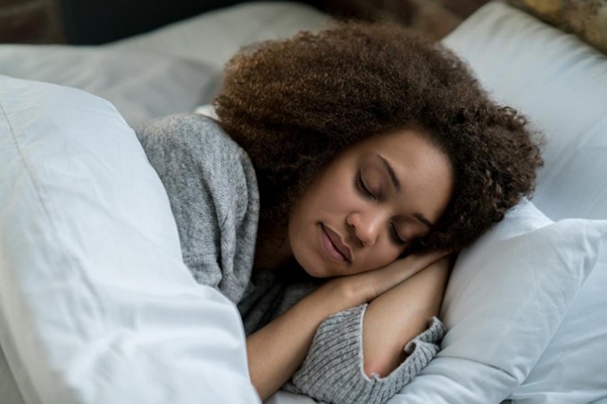 Woman is sleeping in a white bed and wearing a gray sweater, with her arms under her head