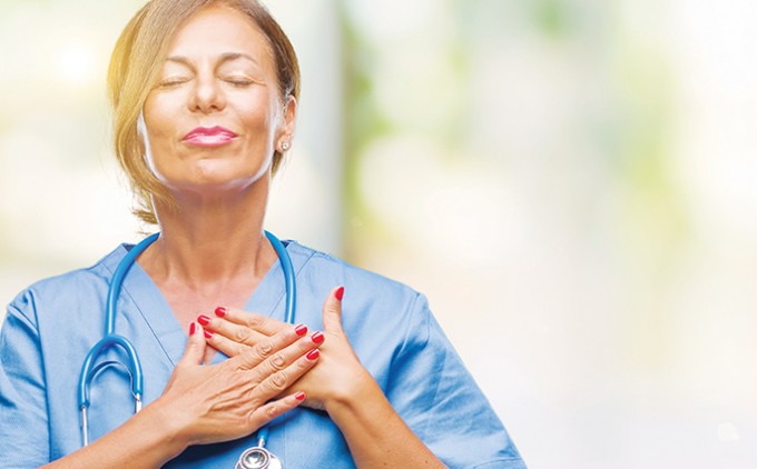 A nurse in scrubs is smiling with her eyes closed and hands on her heart