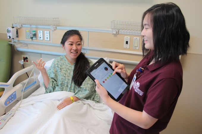 A patient sits upright in a hospital bed talking to a nurse in scrubs at the bedside. The nurse is using a tablet