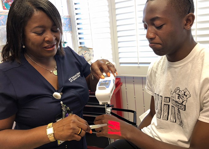 Patient is sitting on a chair with his hand up. Nurse is using a vein finder on his hand and trying to stick in an IV.