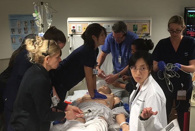 Many nurses and doctors surround a simulation where someone is receiving emergency treatment. Many people are in different roles, like one person is giving CPR