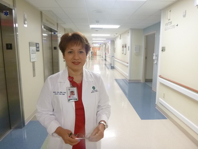 Registered Nurse Nohemy Sam wears a white coat and badge. She is standing and smiling with the hall of her unit behind her.