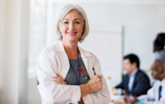 A nurse practitioner is standing and smiling with a white coat on