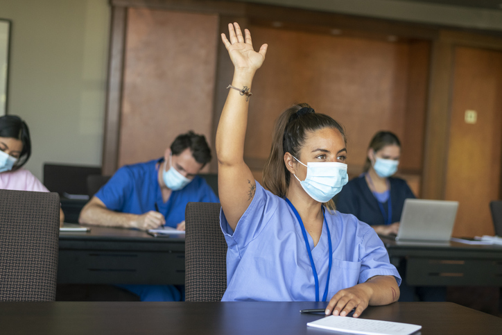 A nurse in a classroom is raising her hand. She and everyone around her is wearing scrubs and look at notebooks or computers