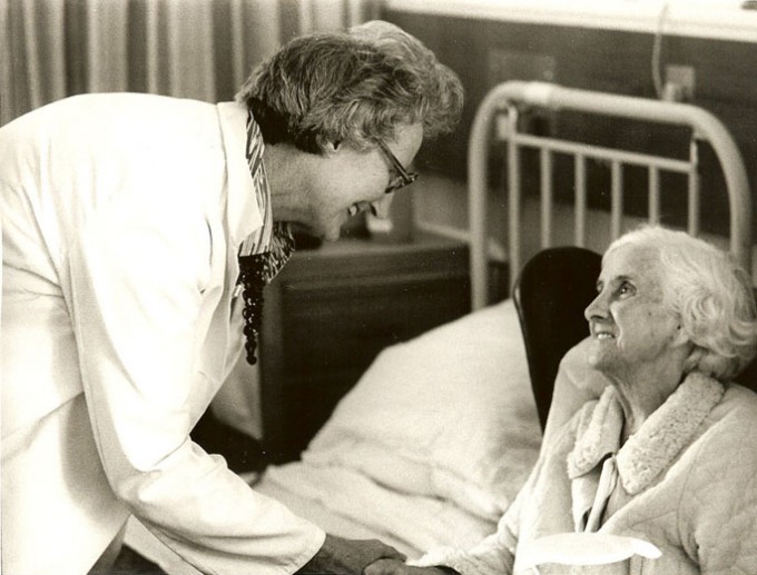 Cicely Saunders bends over to talk to her older, female patient lying in a hospital bed