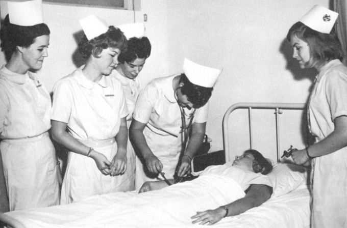 Multiple nurses stand around a patient in a bed. They are wearing white dresses and nursing caps