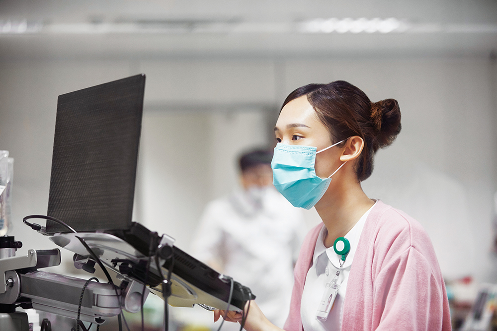 Female nurse wearing a mask, scrubs, and badge, sits in front of a computer that she is looking at