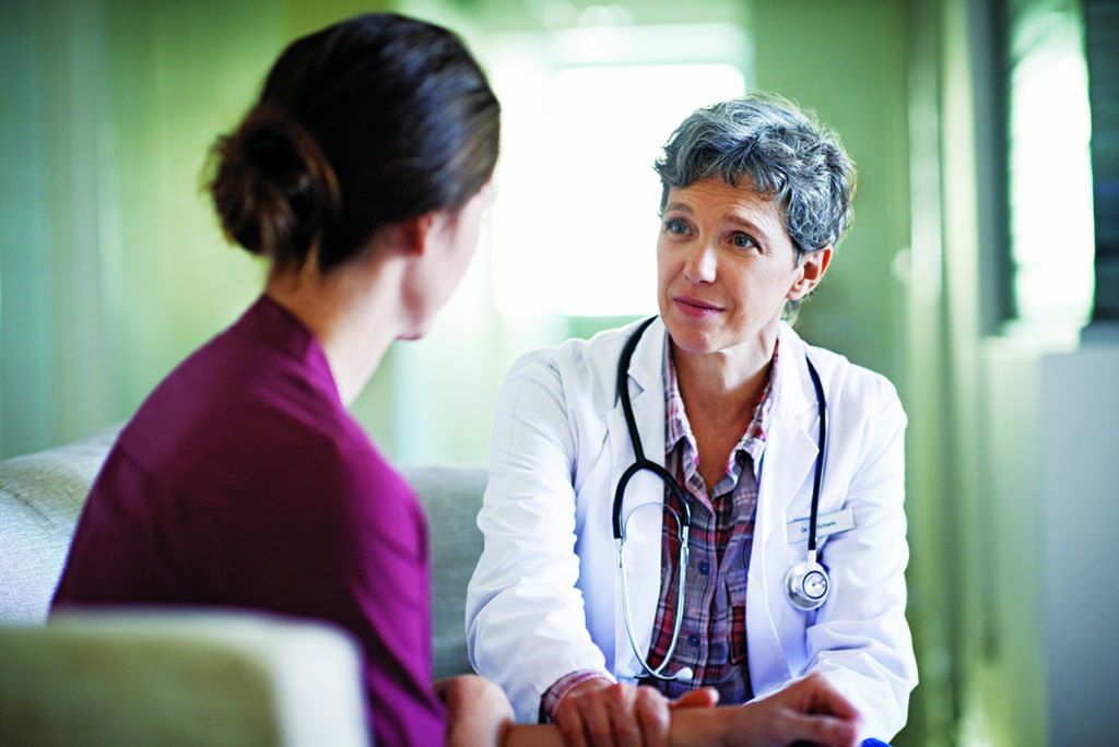 A nurse in a white coat sits across from a young woman, whose back is towards us. They are talking.