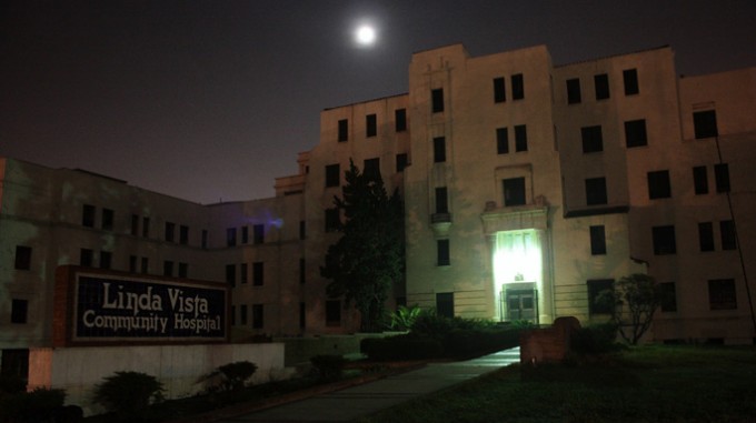 Linda Vista hospital in the eerie darkness of night, only lit by a front door light and the moon in the background. It is a yellow building with 5 floors and dark windows