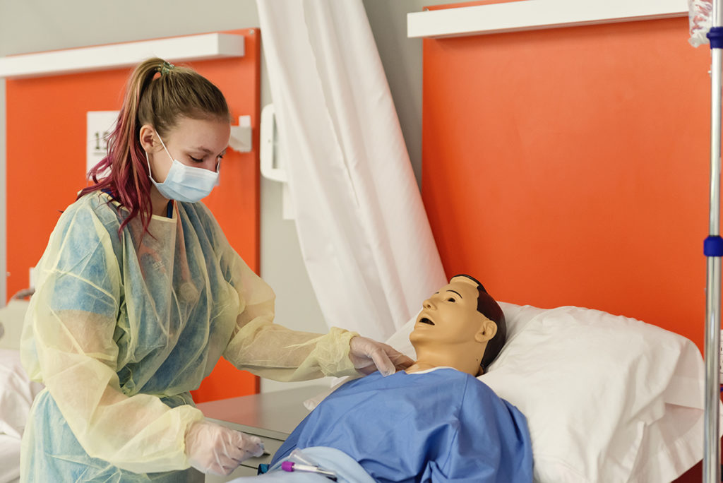Nursing student in isolation gown and mask is listening with stethoscope to a male simulation patient